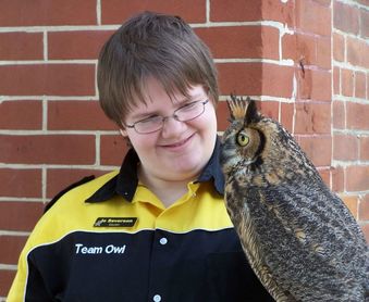 Photo of Jo Severson wearing the owl center uniform and holding Ruby the great horned owl against a brick background