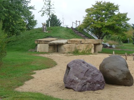 Photo of the natural playground including two climbing rocks, sand pit, stone tunnel, and in the distance is the embankment slide and owl statue on the hill.